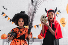 Creepy African American Boy In Halloween Costume Holding Skull Near Spooky Sister With Broom And Carved Pumpkin
