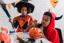 African American Boy In Halloween Costume Carving Pumpkin Near Blurred Sister With Broom
