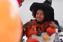 Happy African American Girl In Witch Costume Looking At Brother Carving Pumpkin