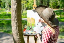 Cheerful Woman Artist At The Plein Air Painting. She Paints An Oil Painting On The Canvas In The Park. Her Canvas Stands On An Easel. Girl Holds A Brush And Palette In Her Hands. Creative Mood.