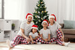 © Syda Productions - family, winter holidays and people concept - happy mother, father and two daughters in santa hats sitting under christmas tree at home