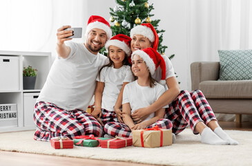 Poster - family, winter holidays and people concept - happy mother, father and two daughters in santa hats sitting under christmas tree at home and taking selfie with smartphone