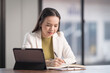 © EduLife Photos - Adult cheerful businesswoman in business outfits writing on notebook with digital table at her working desk with in office