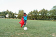 © skif - little boy child in a superhero costume is playing football in the fresh air. happy boy in a red raincoat and a mask runs with a ball on the football field. Outdoor games, dynamic image