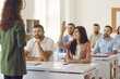 © Studio Romantic - Smiling young woman from student audience raises hand to ask teacher a question. Group of adult people listening to business trainer sitting at tables in classroom during corporate training or seminar