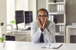 © Studio Romantic - Portrait of smiling super excited overjoyed young black woman in suit sitting at office desk and looking at camera with happy facial expression. Concept of business and career success