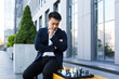 Serious asian man playing chess outside, businessman thinking playing chess sitting on bench near office center
