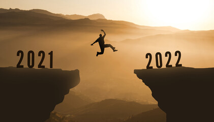Man Silhouette  Jumping from 2021 to 2022 Over Mountain. happy new year 2022.  Sunset Mountains background.  Young guy  Jumps between two Cliffs or two years concept 