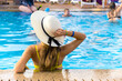© bilanol - Back view of young woman with long hair wearing straw hat relaxing in summer in hotel swimming pool with blue water on a sunny day.