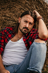 Poster - Young handsome Serbian male in a flannel shirt and ripped jeans sitting leaned on a haystack