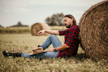 Poster - Young handsome Serbian male in a flannel shirt and ripped jeans sitting leaned on a haystack