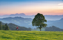Nice Senior Woman On Electric Mountain Bike At Sunset In The Mountains Of Bregenz Forest, Vorarlberg, Austria With Saentis Summit In Switzerland In The Background