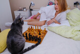 Fototapeta Koty - Beautiful woman playing chess in a bedroom with a cat