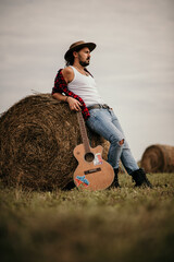 Wall Mural - Young handsome Serbian male in a flannel shirt leaned on a haystack holding his guitar