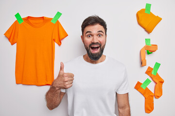 Wall Mural - Positive bearded European man in casual t shirt keeps thumb up poses against white studio background with clothes items recommends something being in good mood glad after cleaning out wardrobe.