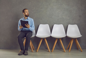 Stressed caucasian man feeling fear before job interview sitting on chair holding clipboard with CV resume and looking aside. Candidate await his turn. Shock people emotion and work search concept