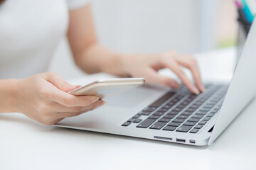 Closeup hand of young woman working laptop computer and reading smartphone on internet online on desk at home, freelance girl using phone with social media, business and communication concept.