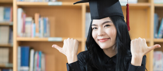 Wall Mural - Portrait of a young Asian university female graduate in graduation gown and mortarboard looking at camera celebrates diploma degree with book self in background
