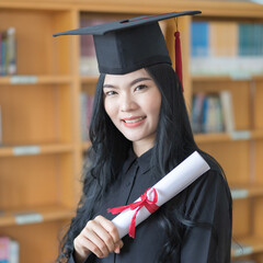 Wall Mural - Portrait of a young Asian university female graduate in graduation gown and mortarboard celebrates diploma degree with book self in background