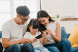 © tirachard - Happy Asian family dad, mom and daughter playing funny game online on smartphone sitting sofa in room at house. Self-isolation, stay at home, social distancing, quarantine for coronavirus prevention.