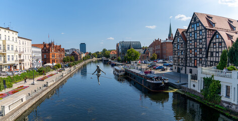 Wall Mural - view of the historic waterfront warehouses and buildings on the Brda River in downtown Bygdoszcz with the Crossing the River sculpture in the foreground