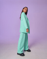 Wall Mural - Elegant woman in mint costume blazer and pants and black top with stripes and boots standing and posing at purple background, Full length fashion portrait.