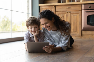 Wall Mural - Happy young hispanic mother using digital tablet with laughing little preschool kid son, lying together on warm wooden floor in kitchen, watching funny cartoons or video content in social network.