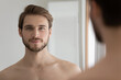 © fizkes - Head shot portrait young handsome good-looking groomed shirtless serious man standing in front of mirror looking at camera feels confident. Male cosmetics and beauty, morning routine, skincare concept