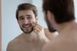 Young man looking in mirror touches face with pimple on cheekbone feels disgruntled. Anti-acne skincare cosmetics treatment, hormonal imbalance, stress, diet, skin problem, freckle appearance concept