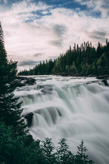 Sticker - Vertical shot of a beautifulwaterfall surrounded with dense green trees under a cloudy sky