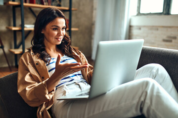 Wall Mural - A woman is sitting on the couch with a laptop in the living room at home. Video conference, work from home. A woman with a laptop communicates, rests and has fun.