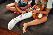 Little Girl With A Broken Leg On The Couch. Two Sisters Draw With Felt-tip Pens On A Plaster Bandage. Children Have Fun And Play On The Living Room Couch.