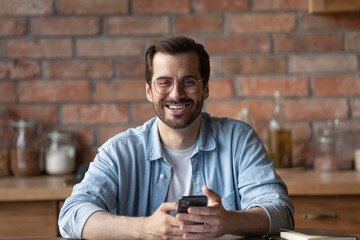 Wall Mural - Head shot portrait smiling man in glasses holding smartphone, sitting at home, happy bearded businessman freelancer or student looking at camera, having fun with mobile device, chatting online