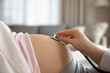 Prenatal health care. Close up of young pregnant woman visiting doctor at antenatal clinic. Medic gynecologist examine fetal heartbeat rate moving stethoscope by naked baby bump of expectant mother