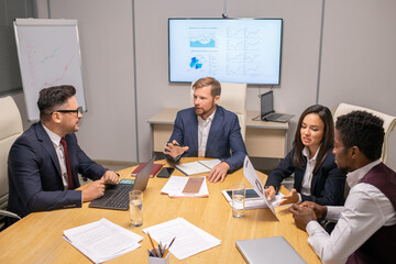 Poster - Two businessmen consulting about speech points while two intercultural colleagues discussing papers by table in boardroom