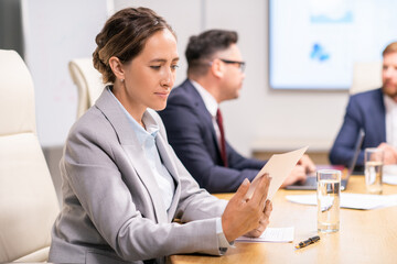 Wall Mural - Young elegant businesswoman with paper sitting by table against male colleagues