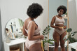 Young woman in underwear measuring her waist with tape in front of mirror