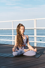 Wall Mural - Contemporary sportswoman sitting in pose of lotus on pier against blue sky and water