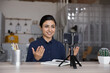 Smiling Indian young woman blogger recording video, using smartphone on tripod, sitting at home office desk, mentor coach or speaker explaining, shooting webinar or training on mobile device
