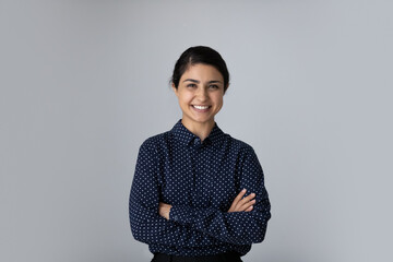 Head shot portrait smiling young Indian woman with arms crossed standing on grey studio background isolated, confident successful businesswoman or student with folded hands looking at camera