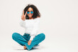 Beautiful black woman with afro curls hairstyle.Smiling model in sweater and trendy jeans clothes. Sexy carefree female sitting on white background in studio. Tanned and cheerful  in sunglasses