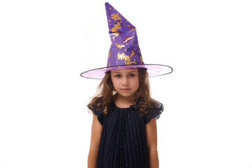 Wall Mural - Portrait of gorgeous confident little girl wearing a wizard hat and dressed in stylish carnival dress, looking at camera posing with crossed arms against white background, copy space.