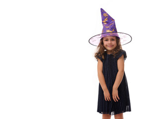 Wall Mural - Portrait of pretty little girl wearing a wizard hat and dressed in stylish carnival dress, looking at camera posing with crossed arms against white background. Halloween concept with copy space
