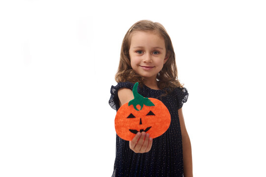Isolated portrait on white background with copy space of adorable little girl, 4 years old pretty kid, holding a homemade felt-cut pumpkin, symbol of the Halloween party, and showing it to the camera