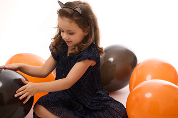 Wall Mural - Adorable baby girl, 4 years old child wearing an evening dress and hoop with cat ears, having fun playing with bright orange and black balloons on white background with copy space. Halloween concept