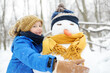 Little boy building snowman in snowy park. Child embracing snowman wearing hat and scarf. Active outdoors leisure with children in winter. Kid during stroll in a snowy winter park