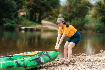 A young woman with diligence pulls a kayak to the river bank with two hands. The concept of active recreation and kayaking