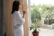 © Andrii Lysenko - morning hot coffee in the hands of a Caucasian brunette standing in the bedroom by the window. A girl in a shirt drinks a hot drink and is resting looking outside the profile view.