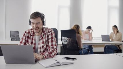 excited young man in headphones working on laptop in office, online music app