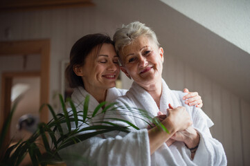 Wall Mural - Happy senior mother in bathrobe with adult daughter hugging indoors at home, selfcare concept.